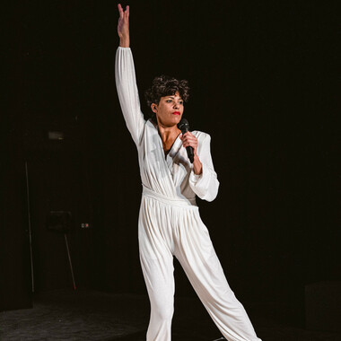 Luanda Casella in a chic white jumpsuit. She holds up one hand and a microphone in the other. Her left leg makes a sidestep.