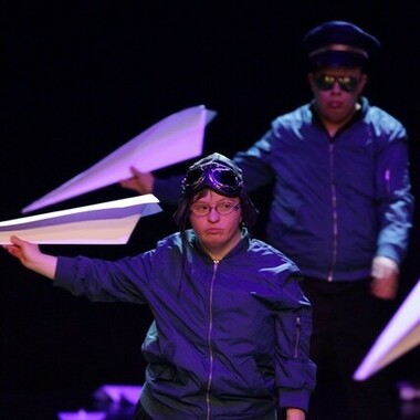 Zwei Performer*innen in Pilot*innen-Kostümen mit jeweils einem großen Papierflieger in der Hand.