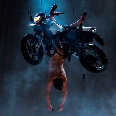 A motorbike floats in the air. A performer hangs upside down from the motorbike.