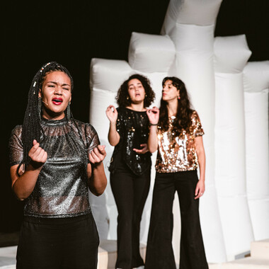 Eine Performerin singt und wird im Hintergrund von zwei weiteren Performerinnen begleitet.