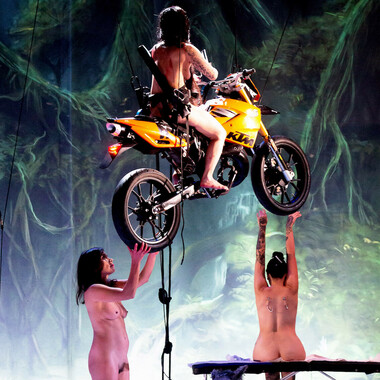 A naked woman on a flying motorbike. Another performer stands under the motorbike and a third sits on a bench in the background.