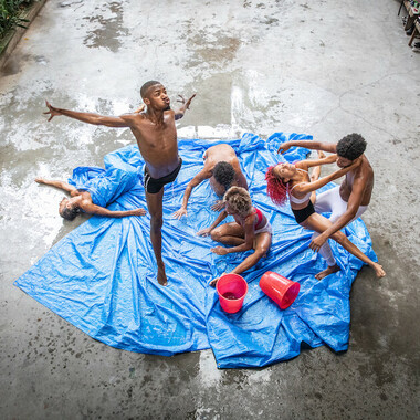 A large canvas on which two buckets of water have been dumped. Six people moving on it. Some dance and jump, others sit or lie down. 