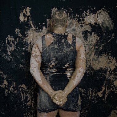 Mira Hamdi from behind. She is standing in front of a black wall with traces of clay on it. Her hands are folded behind her back.