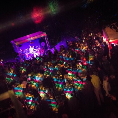Eine Konzertsituation bei Nacht. Das Foto wurde von oben geschossen und zeigt eine Menschenmenge und eine Bühne, die von unterschiedlich farbigen Scheinwerfern beleuchtet werden.