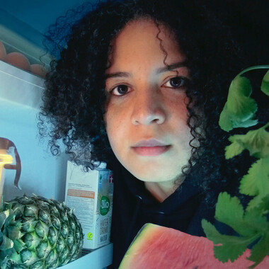 Close-up of the performer from inside a refrigerator. She looks into the fridge and directly into the camera. Her head is surrounded by food: Herbs, oat drink, pineapple, watermelon.