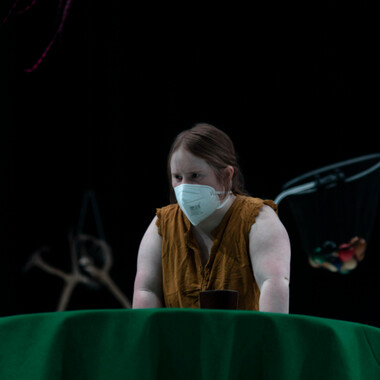 Eine Performerin stützt sich konzentriert mit den Händen auf einem runden Tisch ab. Auf dem Tisch ist eine grüne Tischdecke aus festem Stoff.