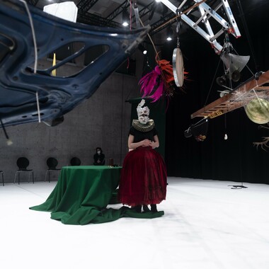 A performer wears a costume with a wide, long skirt and a large fan collar. Her face is masked. Behind her is a round table with a dark green tablecloth. A construct of ladders hangs from the ceiling.