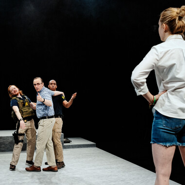 Image description by the editorial staff of Festival Theaterformen. Self-description of the depicted persons follows. / In the background on the left, three people are standing and looking forward. Two of them are wearing clothes with the inscription FBI. On the right, one person stands in the foreground with their hands on their hips. They are wearing a white shirt and jean shorts and are looking in the direction of the other three people. 