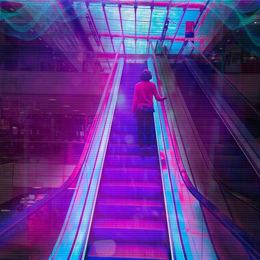 Image description by the editorial staff of Festival Theaterformen. Self-description of the depicted persons follows. / A person with headphones centered on a moving escalator in a shopping mall. The person is standing with their back to the camera, and they are touching the escalator handrail with their right hand. There is a filter over the photo in shades of pink and blue.