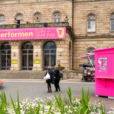 Infobox auf dem Vorplatz vor dem Großen Haus: Eine neonpinke Holzhütte, nach vorne hin geöffnet, im Hintergrund das Große Haus mit großem pinkem Festivalbanner.