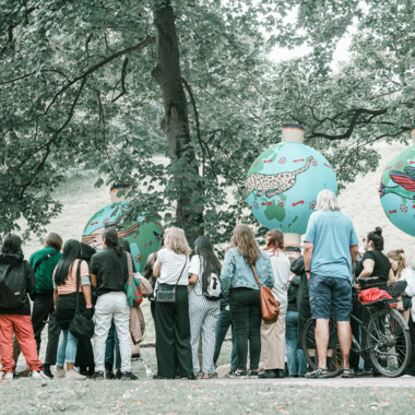 Eine Menschengruppe von etwa 20 Personen steht vor itaakás, die in den Bäumen des Theaterparks hängen.