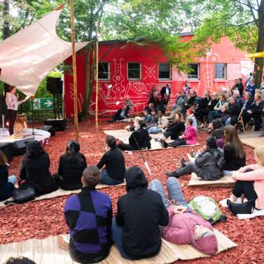 Festivalzentrum am Gartenhaus Haeckel: Der Boden ist mit rotem Hackschnitzel belegt, auf dem Matten liegen, darauf sitzen Menschen. Links eine Bühne mit darüber gespanntem Tarp.