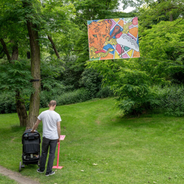 A large colourful banner with hand-painted drawings stretched between trees at all four corners.  A person with a pram stands under a large colourful banner with hand-painted drawings stretched between trees at all four corners and reads a description.