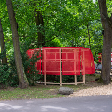 A photo of the installation Beneath the Mud of the Ground I: It shows a wooden construction with transparent red fabric hanging from the top. In the middle are trees and bushes. The construction is surrounded by trees.