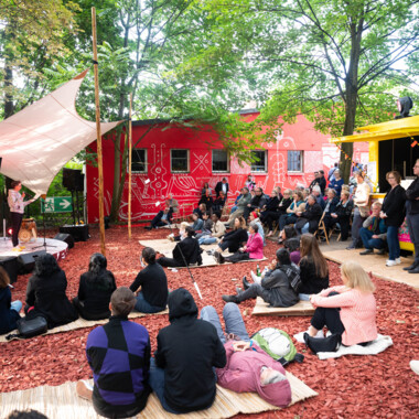 Festival centre at Gartenhaus Haeckel: The floor is covered with red wood chips, with mats and people sitting on them. On the left, a stage with a tarp stretched over it.