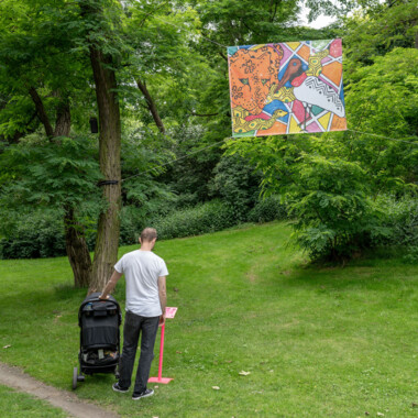 Eine Person mit Kinderwagen steht unter einem großen bunten Banner mit handgemalten Zeichnungen, das an allen vier Ecken zwischen Bäumen gespannt ist und liest sich eine Beschreibung durch.