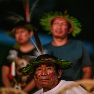 Drei Personen der Baniwa Gemeinschaft, ihre Gesichter sind bemalt, sie tragen Kopfbedeckungen aus Planzen.