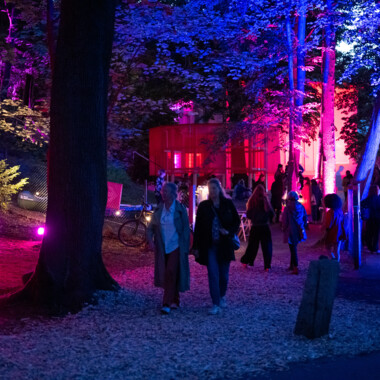 Festival centre at Gartenhaus Haeckel in the evening light. The trees are illuminated in blue and red, people are walking on paths.
